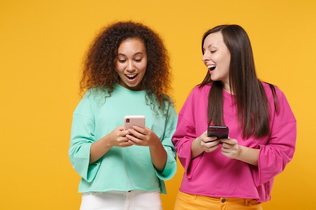 Dos amigas emocionadas chicas afroamericanas europeas vestidas de verde rosa posando aisladas en un fondo amarillo. Concepto de estilo de vida de las personas. Simulacros de espacio de copia. Usando el teléfono móvil, escribiendo un mensaje.