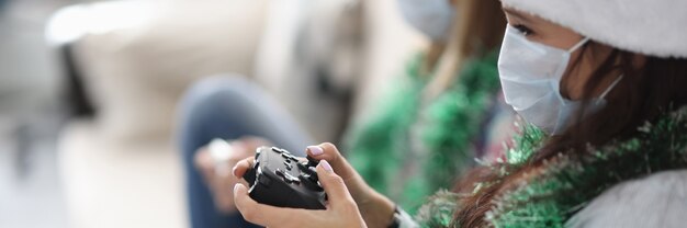 Dos amigas están sentadas en el sofá con joysticks de juego con sombreros de santa claus y medi ...