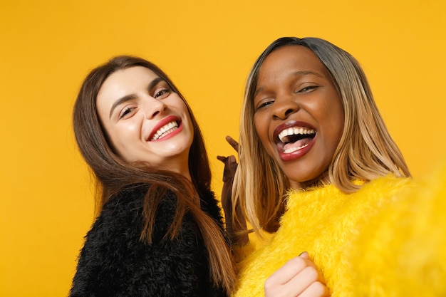 Dos amigas europeas y afroamericanas vestidas de amarillo negro haciendo selfie posando aisladas en un retrato de estudio de fondo de pared naranja brillante. Concepto de estilo de vida de las personas. Simulacros de espacio de copia