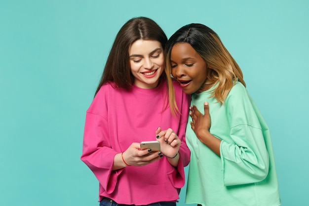 Dos amigas europeas y afroamericanas vestidas de verde rosa sostienen un celular en la mano aislado en un fondo azul turquesa de la pared, retrato de estudio. Concepto de estilo de vida de las personas. Simulacros de espacio de copia.