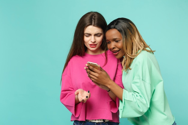 Dos amigas europeas y afroamericanas vestidas de verde rosa sostienen un celular en la mano aislado en un fondo azul turquesa de la pared, retrato de estudio. Concepto de estilo de vida de las personas. Simulacros de espacio de copia.