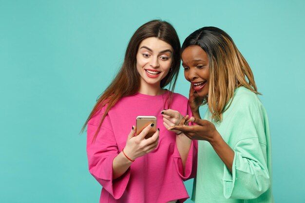 Dos amigas europeas y afroamericanas vestidas de verde rosa sostienen un celular en la mano aislado en un fondo azul turquesa de la pared, retrato de estudio. Concepto de estilo de vida de las personas. Simulacros de espacio de copia.