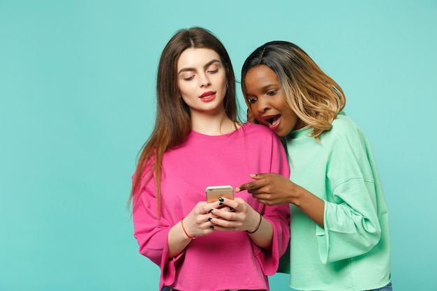 Dos amigas europeas y afroamericanas vestidas de verde rosa sostienen un celular en la mano aislado en un fondo azul turquesa de la pared, retrato de estudio. Concepto de estilo de vida de las personas. Simulacros de espacio de copia.