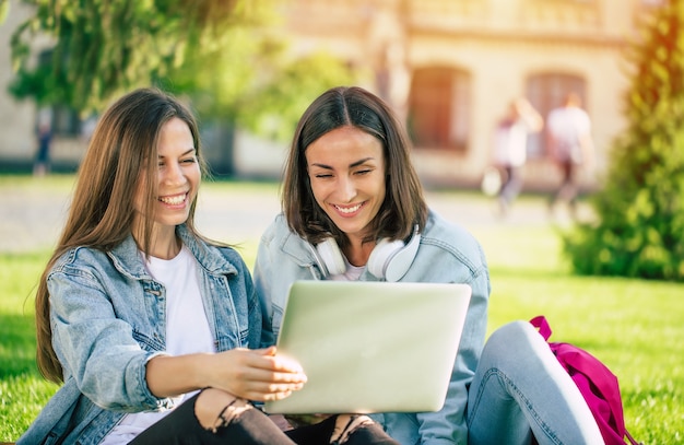 Dos amigas feliz hermosa joven estudiante en ropa casual de mezclilla se están relajando en el parque universitario con la computadora portátil y el teléfono inteligente por la universidad y tomando café