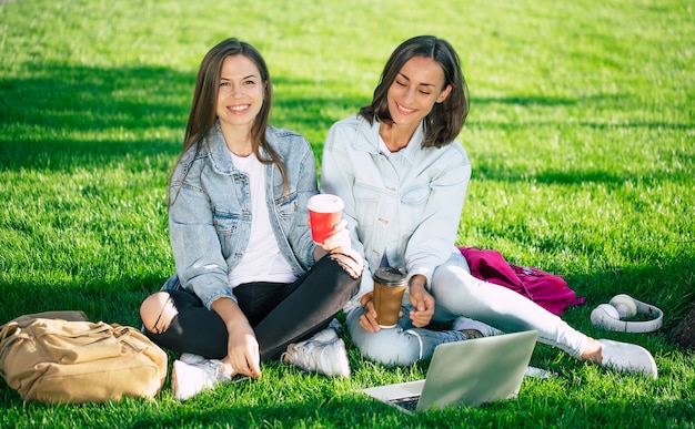 Dos amigas feliz hermosa joven estudiante en ropa casual de mezclilla se están relajando en el parque universitario con la computadora portátil y el teléfono inteligente por la universidad y tomando café