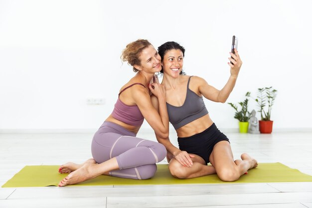 Foto dos amigas de fitness sentadas juntas en la alfombra y tomando selfies en el teléfono en el interior