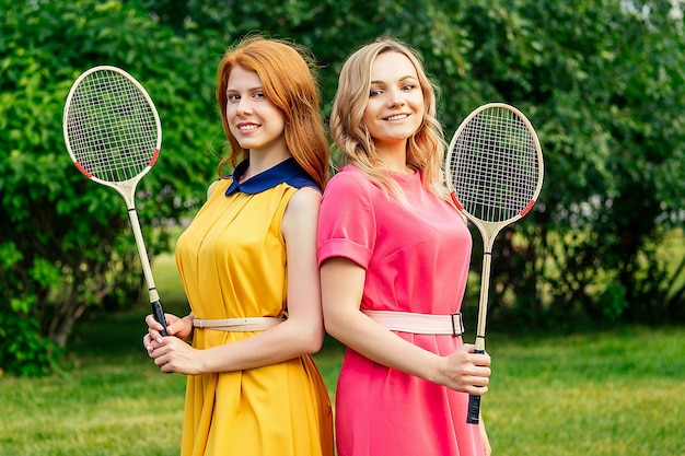 Dos amigas hermosa joven pelirroja pelirroja irlandesa con un vestido amarillo y una persona femenina rubia europea con un vestido rosa sosteniendo una raqueta de bádminton de tenis en el parque de verano