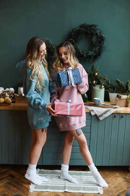 Dos amigas hermosas mujeres en sudaderas con capucha de moda se dan cajas de regalo de Navidad en el interior decorado para las vacaciones de año nuevo
