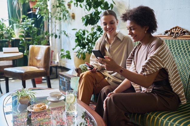 Dos amigas interculturales sonrientes viendo un video gracioso en su teléfono inteligente