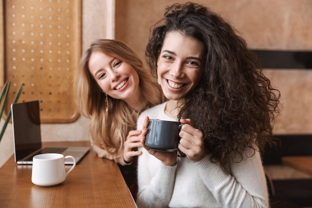 Dos amigas jóvenes alegres sentados en el café en el interior, tomando una taza de café