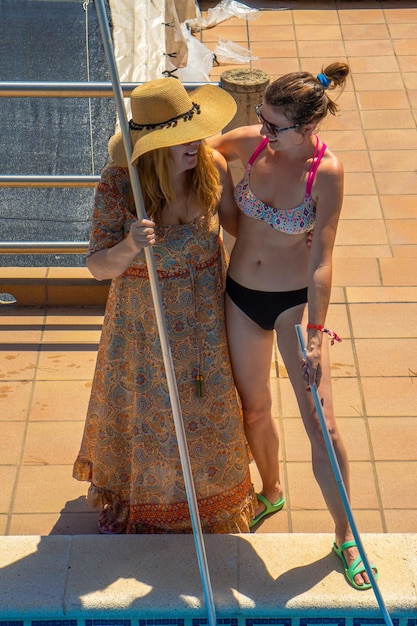 Dos amigas lesbianas pareja abrazándose mirándose alegremente antes de limpiar una piscina con vestido de verano y bikini sosteniendo los postes de limpieza en el fondo de la piscina