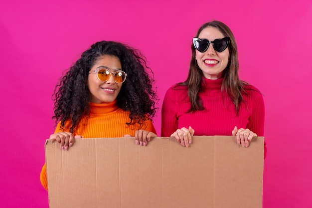 Dos amigas multiétnicas sonrientes sosteniendo un cartel de cartón sobre un fondo rosa