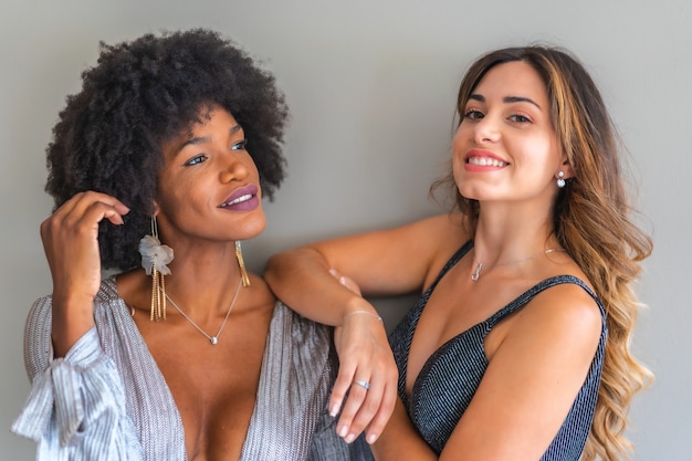 Dos amigas multiétnicas en vestidos de gala listas para salir de noche a la fiesta exclusiva. Vestidos de noche grises y azules, chica rubia caucásica, chica negra con pelo afro