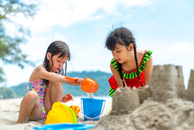 Dos amigas niñas asiáticas pequeñas sentadas en una playa tropical jugando a la arena con juguetes de playa juntas en las vacaciones de verano niños felices niño disfruta y diversión actividad al aire libre estilo de vida en vacaciones de playa