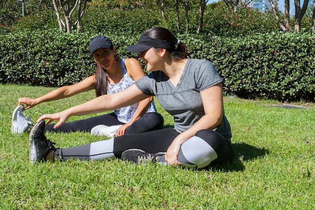 Dos amigas en el parque estirando las piernas para comenzar su entrenamiento mientras se divierten