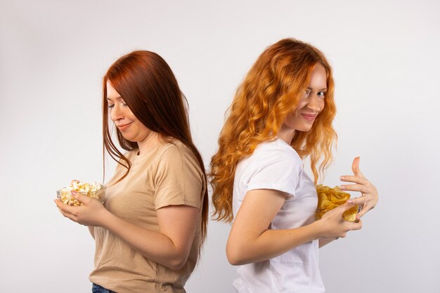 Dos amigas posando sobre una pared blanca escondiendo cuencos con palomitas de maíz y papas fritas