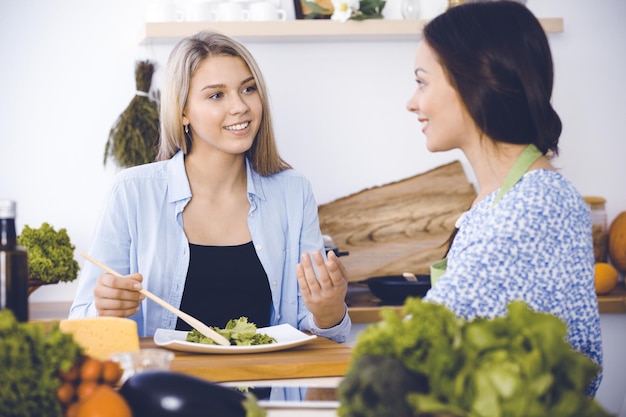 Dos amigas prueban nuevas recetas de deliciosas ensaladas frescas mientras se sientan en la mesa de la cocina Concepto de cocina vegetariana