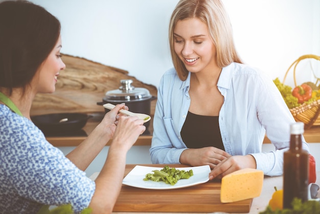 Dos amigas prueban nuevas recetas de deliciosas ensaladas frescas mientras se sientan en la mesa de la cocina Concepto de cocina vegetariana