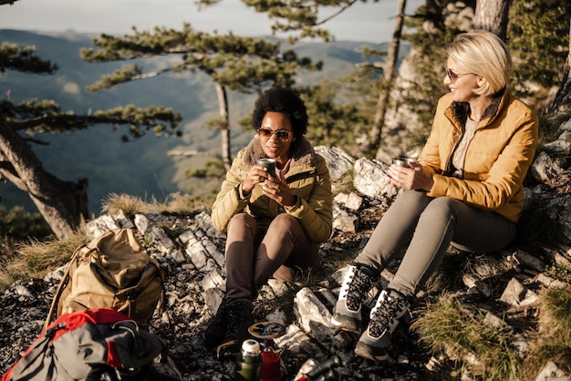 Dos amigas de raza mixta toman un descanso para el té a mitad de su caminata en las montañas en un hermoso día soleado de otoño.