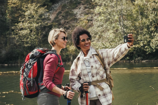 Dos amigas de raza mixta tomándose selfies junto a un río durante una caminata por la montaña.