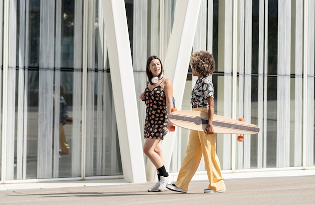 Dos amigas en ropa casual paseando por la ciudad, amigas multietincas, con una patineta