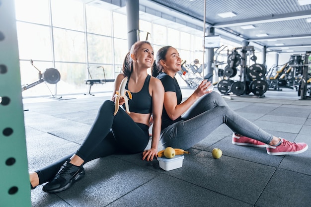 Dos amigas en ropa deportiva están en el gimnasio comiendo frutas y tomando un descanso.