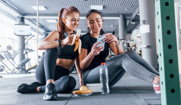 Dos amigas en ropa deportiva están en el gimnasio comiendo frutas y usando el teléfono.