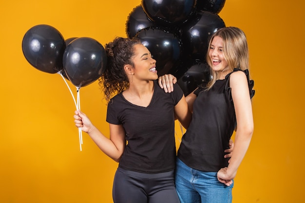 Dos amigas sobre fondo amarillo con globos negros en la fiesta de promoción del viernes negro.