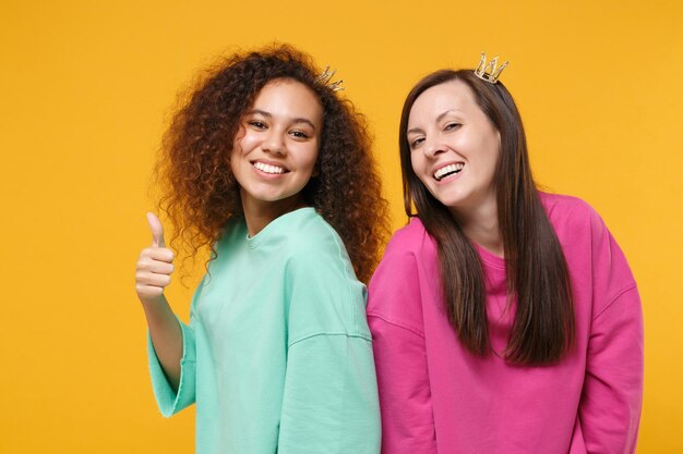 Foto dos amigas sonrientes chicas europeas y afroamericanas vestidas de verde rosa, coronas posando aisladas en el fondo de la pared amarilla. concepto de estilo de vida de las personas. simulacros de espacio de copia. mostrando el pulgar hacia arriba.