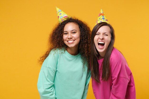 Dos amigas sonrientes gritando chicas europeas y afroamericanas vestidas de verde rosa, sombreros de cumpleaños posando aisladas en un fondo naranja amarillo. Concepto de estilo de vida de las personas. Simulacros de espacio de copia.