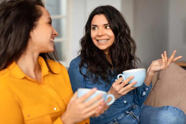 Foto dos amigas sonrientes hablando sentadas con tazas de té en el interior