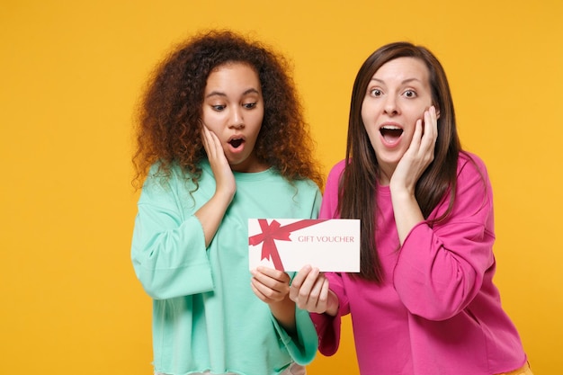 Dos amigas sorprendidas, chicas europeas y afroamericanas vestidas de verde rosa posando aisladas en un fondo naranja amarillo. Concepto de estilo de vida de las personas. Simulacros de espacio de copia. Mantenga el certificado de regalo.