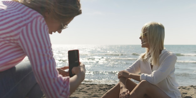 Dos amigas tomando fotos con un smartphone en la playa vacía durante el día de otoño