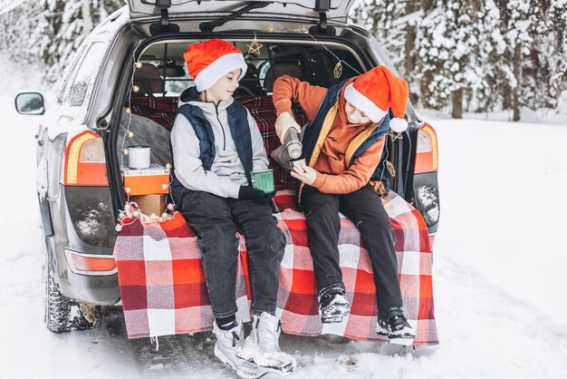 Dos amigos adolescentes muchachos con sombreros rojos de Papá Noel con destellos de fuego de Bengala parados cerca del baúl del auto decorado para Navidad y Año Nuevo en un bosque nevado de invierno Viaje por carretera y viajes locales