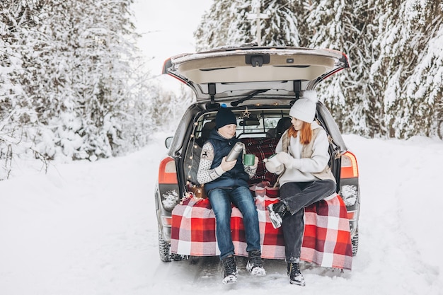 Dos amigos adolescentes niño y niña con termo sentado en el maletero del coche decorado para Navidad Aventura de viaje por carretera y concepto de viaje local Picnic de invierno en el bosque nevado Vacaciones de año nuevo