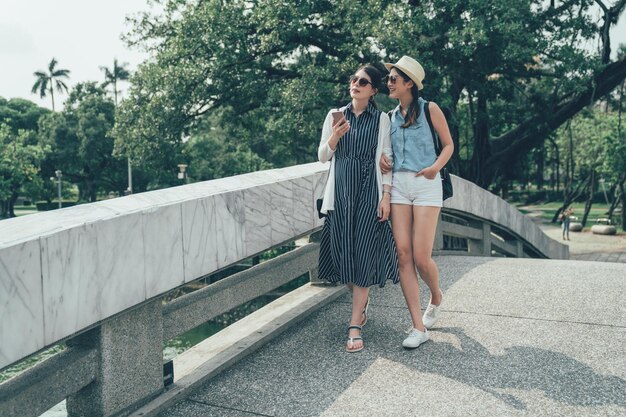 dos amigos asiáticos de cuerpo entero caminando por el puente en el parque tradicional chino en taiwán taichung. chicas jóvenes comunicándose y usando teléfonos móviles junto a la piscina natural. las mujeres viajeras navegan por la aplicación de mapas