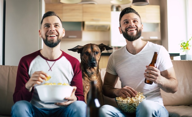 Dos amigos barbudos emocionados felices viendo la televisión o un partido de deporte con el perro mientras están sentados en el sofá en casa el fin de semana