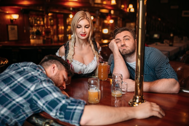 Dos amigos borrachos durmiendo en el mostrador con jarras de cerveza en el pub, bonita camarera