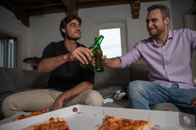 Dos amigos brindan con dos cervezas en un descanso del videojuego comiendo pizza