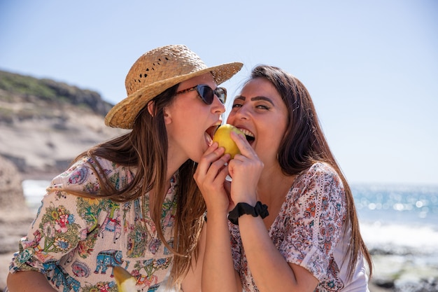 Foto dos amigos comen juntos una manzana en la playa, felices, sonrientes y disfrutando de sus vacaciones.