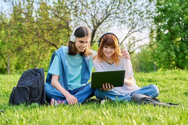 Dos amigos estudiantes chico y chica en auriculares mirando portátil sentado en el césped
