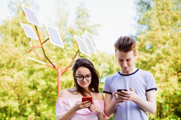 Dos amigos estudiantes usando teléfonos móviles contra paneles solares en el parque