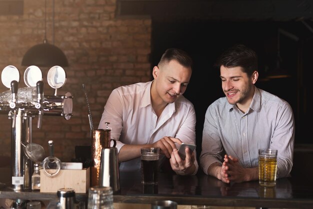 Dos amigos felices disfrutando de cerveza fresca en el pub y navegando por las redes sociales en el móvil. Tecnologías, comunicación en línea y adicción a internet, espacio de copia