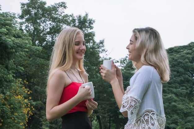 Dos amigos felices hablando sosteniendo una taza de té al aire libre en un parque