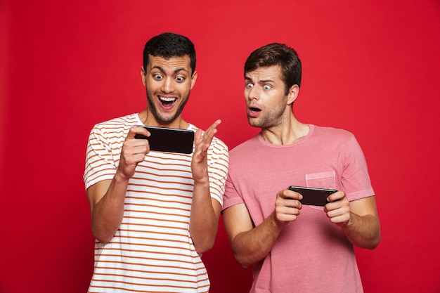 Dos amigos hombres emocionados que se encuentran aisladas sobre la pared roja, jugando juegos en teléfonos móviles