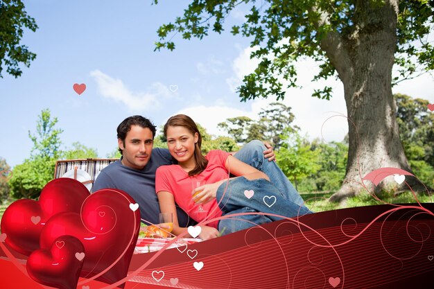 Dos amigos mirando hacia adelante mientras sostienen vasos mientras hacen un picnic contra el diseño del corazón de San Valentín