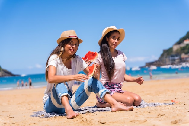 Dos amigos en verano en la playa sonriendo comiendo una sandía con el mar de fondo sentados en la arena