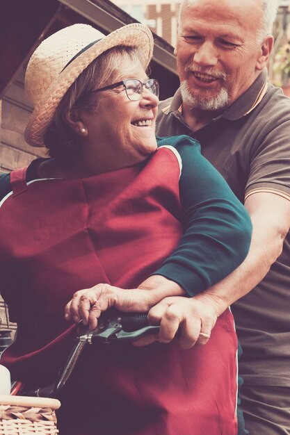 Foto dos ancianos caucásicos, hombres y mujeres mayores, se divierten y disfrutan del amor juntos jugando y koning al aire libre con una bicicleta y una casa de madera en el fondo, una feliz pareja madura sonríe y ríe