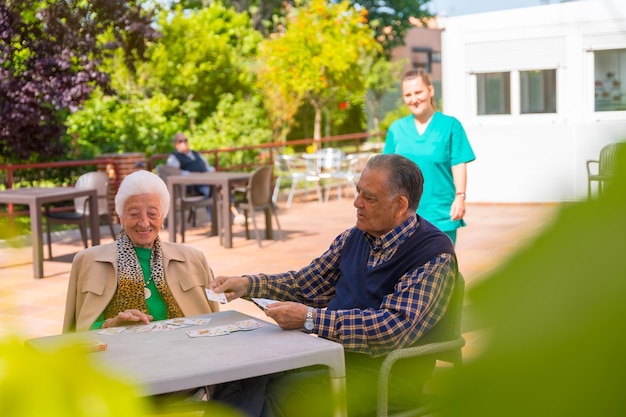 Foto dos ancianos con la enfermera en el jardín de un hogar de ancianos o de retiro jugando a las cartas en una mañana de verano