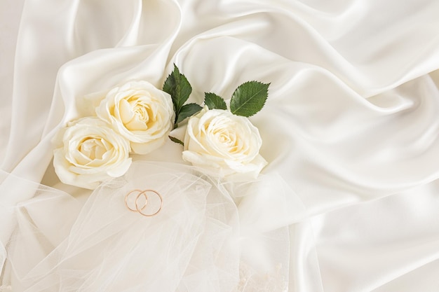 Dos anillos de boda dorados sobre un fondo beige satinado con rosas de té vivas y parte del velo de la novia Diseño elegante para el diseño Fondo de boda